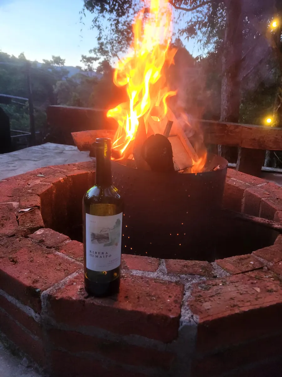 Fogo de chão aceso ao entardecer, com garrafa de vinho sobre a mureta de tijolos e a mata ao fundo