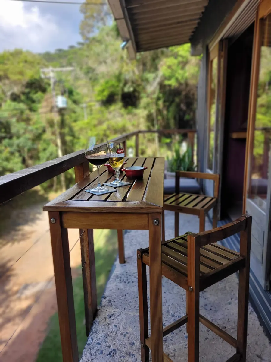Mesa alta e banquetas de madeira na varanda, com taças de vinho e cerveja e vista para a serra